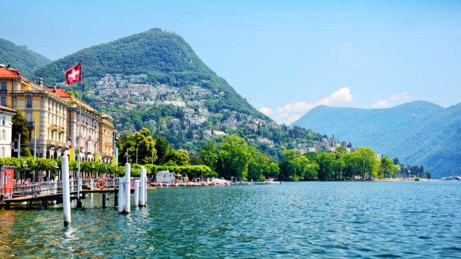 Orla do Lago di Lugano com a bandeira suíça, cercada por montanhas