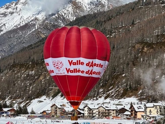 Balão de ar quente com os dizeres Valle d'Aosta sobrevoando um vilarejo nevado, com os Alpes ao fundo